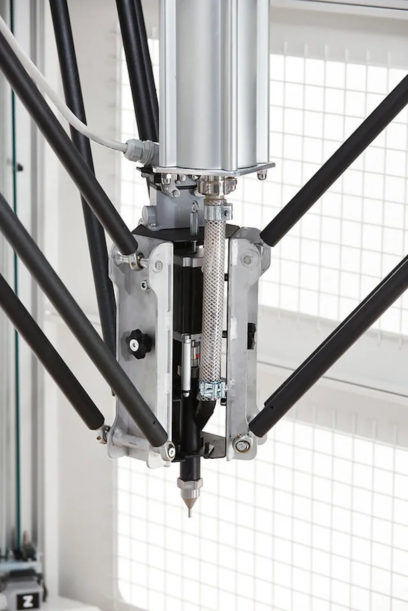 Delta Wasp 3MT 3D printer close-up, showing the extrusion mechanism with black rods and metal components against a grid background.