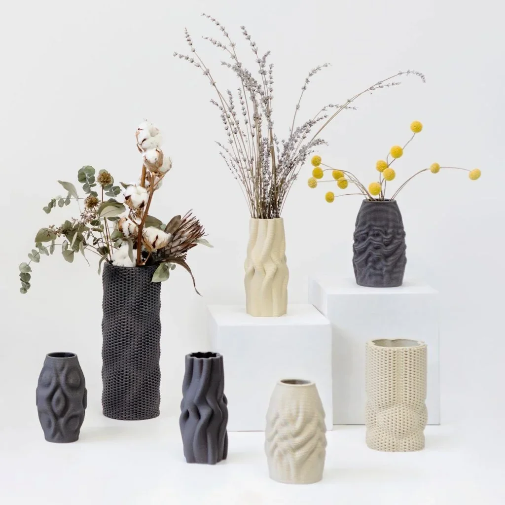 3D-printed vases of varied designs created by Delta WASP 3MT, some filled with flowers, displayed on white pedestals in a minimalistic setting.