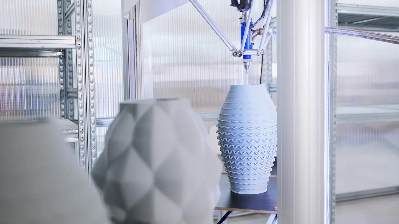 Delta Wasp 40100 Clay 3D printer creating intricate ceramic vase, adjacent to textured finished pots. Industrial setting with metal shelves.