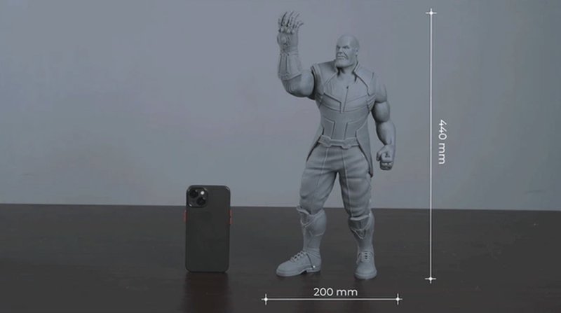 A 3D-printed sculpture, created by the emake3d Galaxy 1, stands next to a smartphone for scale comparison on a dark surface. The sculpture, approximately 440 mm tall and 200 mm wide, depicts a muscular figure with detailed anatomical features. It showcases intricate design elements such as segmented armor and a raised hand, highlighting the precision and capability of the 3D printer. The smartphone, positioned beside the sculpture, emphasizes the model’s size, illustrating the printer’s high-volume capacity. The background is a plain, light-colored wall that creates a neutral setting, focusing attention on the 3D print output.