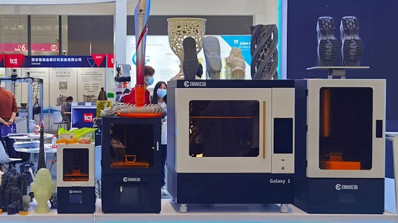emake3d Galaxy 1 3D printer setup with various printed models, including large detailed sculptures and footwear, on display at an exhibition booth.