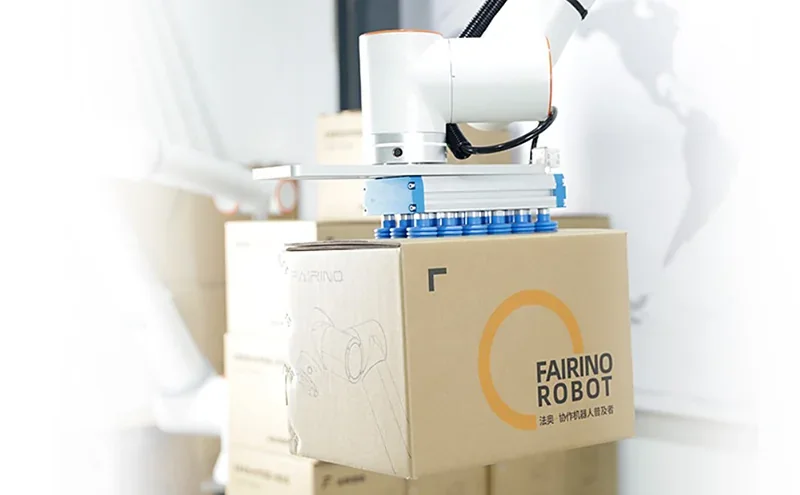 Fairino FR3 robot handling box with blue suction cups, Fairino branding visible, set in industrial area with stacked cardboard boxes.
