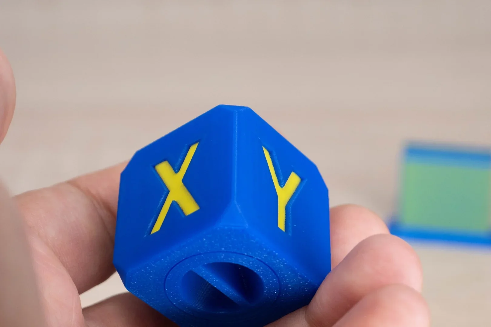 Blue 3D-printed cube with yellow "X" and "Y" text created by FlashForge AD5X, held in hand.