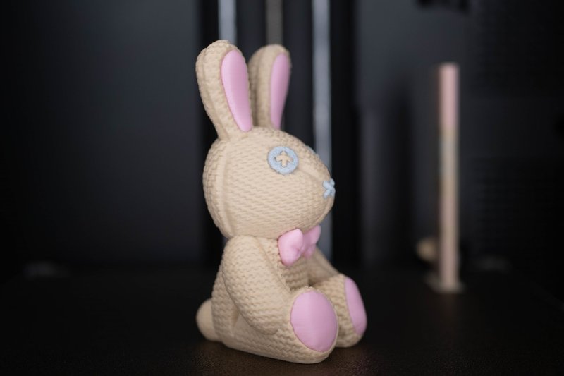 A 3D-printed bunny figurine created by the Flashforge AD5X is showcased with intricate details. The bunny has a textured, beige knitted pattern, adding a realistic fabric-like appearance. Its ears, paw pads, and bow tie are accented in soft pink, contrasting with the beige body. The eyes and nose are designed with a stitched, button-like aesthetic using a pale blue color. The setting is a minimalist black backdrop, which emphasizes the craftsmanship and color details on the bunny. The Flashforge AD5X's capability to produce finely detailed and multi-colored objects is evident in this high-quality print.