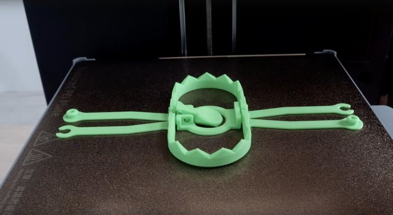 A green plastic 3D-printed object resembling a bear trap is displayed on the build plate of a Flashforge Adventurer 5X 3D printer. The object has a circular base with jagged edges and extended lever arms on both sides, showcasing the printer's precision in creating intricate details. The build plate texture and the Flashforge branding are visible on the left side. The setting appears to be an indoor workspace with the printer's interior partially in view, emphasizing the machine's application in creating complex designs with consistent structural accuracy.