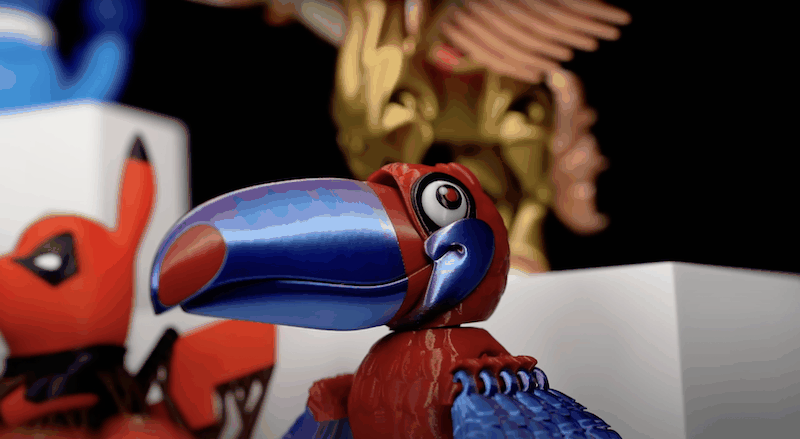 A variety of detailed 3D printed models in vibrant colors showcase the capabilities of the Flashforge Adventurer 5X 3D printer. In the foreground, a large parrot model features a gradient of blue to purple on its beak, with red body details and intricately designed feathers, demonstrating the precision of multicolor printing. Behind it, other models include a highly detailed red and black figure and a gold, winged structure, highlighting the printer’s ability to handle intricate designs and multiple colors. The setting is a display environment with minimalistic white platforms, drawing focus to the vibrant models, indicative of the printer's exceptional output quality.