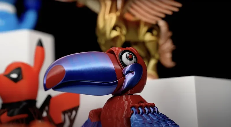 3D-printed colorful toucan figure, vibrant red and blue hues, possibly crafted by Flashforge AD5X; background displays more printed models.
