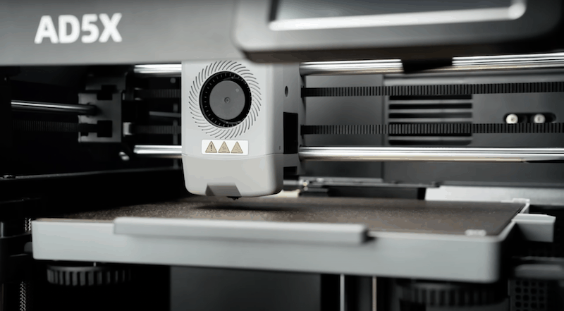 The Flashforge AD5X 3D printer is shown in a close-up view, highlighting its advanced printing head and mechanical components. The machine's branding 