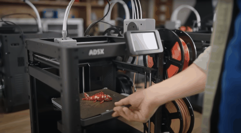 The image showcases the Flashforge AD5X 3D printer prominently in a workshop setting. A black, sturdy frame supports the printer's open design, allowing clear visibility of the printing area. The printer is in the process of creating a detailed, colorful dragon-like figurine, with visible red and yellow segments, using its advanced printing capabilities. Located on the side of the printer is a touch screen interface for user interaction. Behind the touch screen, multiple spools of filament in various colors, including orange and yellow, are mounted on the side, indicating the printer's multi-material capability. The surrounding workshop environment features shelves and other printers, suggesting a professional or educational setting. The branding 