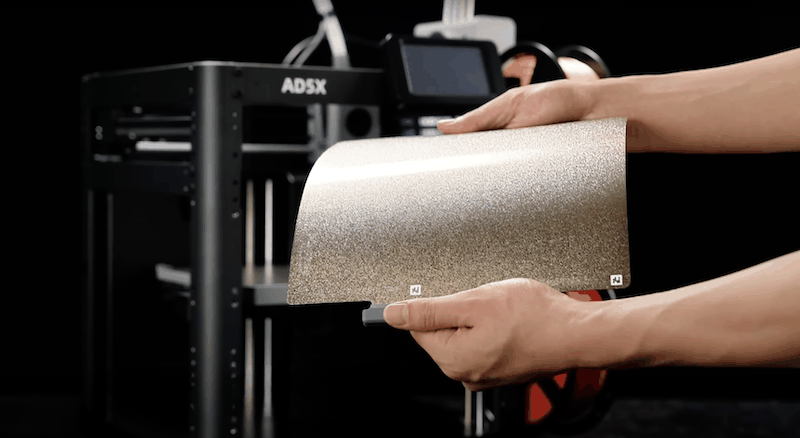 The image showcases a Flashforge AD5X 3D printer in a dimly lit setting with a focus on a textured, flexible build plate held by two hands in the foreground. The printer is visible in the background, featuring a robust frame with a metallic finish and prominent branding displaying 