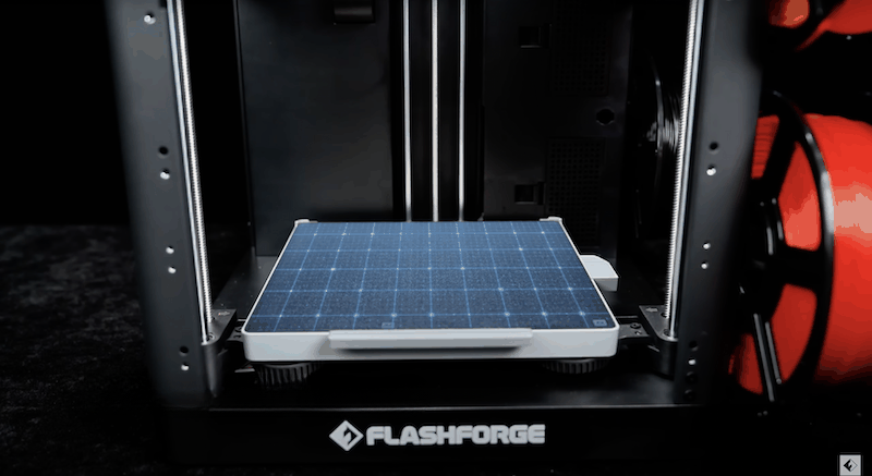 The image shows the Flashforge AD5X 3D printer with the main focus on its build platform, which features a blue grid pattern for precise alignment and measurements. The printer's open frame design reveals dual vertical guide rods on either side for stability and accurate movement of the print head. The Flashforge branding is prominently displayed on the front base of the printer. Spools of red filament are visible to the side, emphasizing the printer’s capability to handle versatile printing materials. The setting is likely a studio or tech workshop, highlighted by a dark background that contrasts with the illuminated printer components.