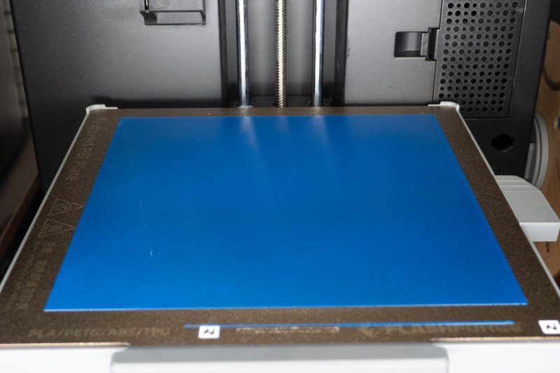 The image showcases the build plate of a 3D printer, featuring a smooth, blue, removable surface designed for ease of model removal. Visible are dual vertical threaded rods, indicative of a stable Z-axis mechanism aimed at precise layer alignment. The surrounding frame highlights detailed markings for material compatibility, suggesting the use of PLA, PETG, ABS, and TPU filaments. The background includes a vented panel typical for cooling or exhaust functions, alongside a partial view of mechanical components indicating robust construction. The image likely focuses on the printer’s technical details rather than branding or overall structure.