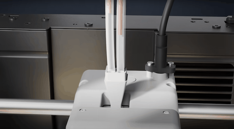 The image shows a close-up view of the Flashforge AD5X 3D printer, focusing on the extruder mechanism. The extruder is encased in a sleek, white housing with visible wiring that feeds filament through a set of guiding tubes. The setup is mounted on a metallic rod suggesting precise movement capabilities, possibly along the X-axis, indicative of the printer's ability to perform detailed print operations. The surrounding area features a black metal casing with ventilation slots, suggesting a well-structured cooling system. The environment appears to be an industrial or workshop setting, emphasizing the printer's professional-grade capabilities. No specific branding is visible on the displayed components.