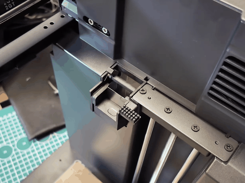 Close-up view of a 3D printer, displaying a part that is possibly a component or accessory related to the Flashforge AD5X. The image focuses on a section of the printer's frame, highlighting a black plastic bracket with a textured surface detail. This bracket is mounted on a metal rod supported by a set of vertical metal bars, secured with several screws. Adjacent to the bracket, a smooth matte-black surface with vent-like cutouts can be seen on the printer's body, indicating potential ventilation for cooling. The setting includes a portion of a workspace with a cutting mat in the background, suggesting a technical or industrial environment where 3D printing assembly or maintenance is performed.