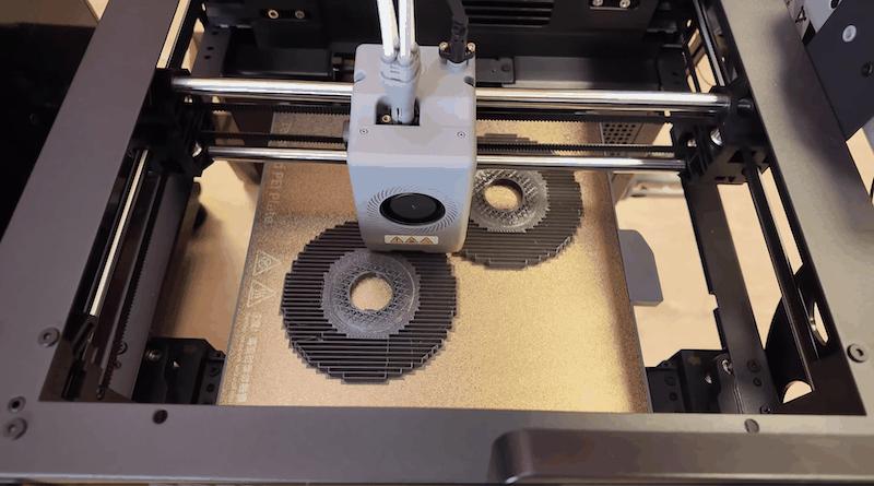 The image shows the Flashforge Adventurer 5X 3D printer in operation. The printer’s build area features a heated print bed, with two circular objects currently being printed, highlighting its active dual-color extruder capabilities. The extruder head is centrally positioned, with white cables attached, suggesting precise filament control. The machine's robust frame and smooth rails ensure stability and accuracy during the printing process. Visible branding includes the Flashforge logo and caution symbols on the extruder, emphasizing safety and brand identity. The printer's modern design and enclosed structure indicate focus on usability and aesthetic integration into various environments.