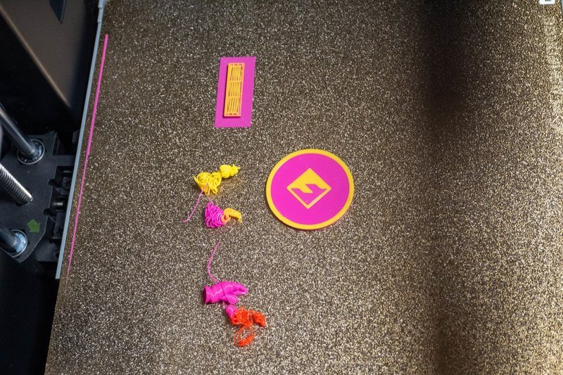 On a textured, metallic print bed inside a 3D printer, various objects created by the Flashforge AD5X are displayed. The objects include a pink and yellow rectangular grid pattern and a circular piece featuring a symbol at the center, outlined in yellow. Additionally, there are colorful abstract shapes in yellow, pink, and orange, resembling small artistic sculptures. The print bed showcases a rough, grainy surface, indicating durability and adhesion properties crucial for successful printing. The surrounding structure of the printer is partially visible, featuring metal rods and structural components, suggesting a robust and precise printing environment. No branding is visible within this close-up setting.