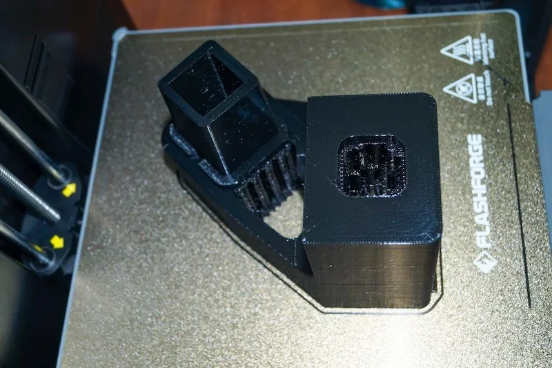 Black 3D printed part on Flashforge Adventurer 5M printer bed, close-up view, showcasing the printer's precision and branding.