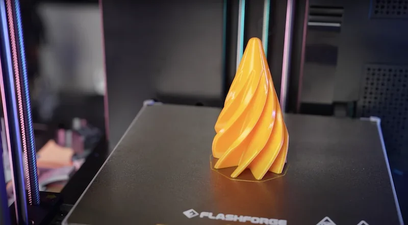 Flashforge Adventurer 5M 3D printer showcasing a yellow spiral cone object on its print bed, with visible Flashforge branding.