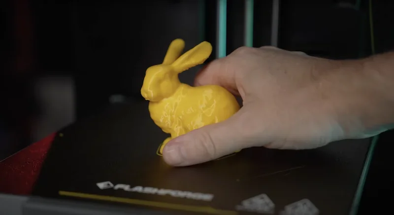 Hand holding a yellow 3D-printed rabbit model on Flashforge Adventurer 5M print bed, showcasing printer's detail and precision. Flashforge logo visible.