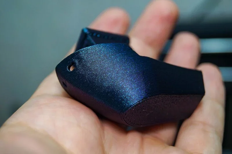 A hand holds a dark blue 3D-printed part, showcasing smooth texture and precision printing by FlashForge Adventurer 5M printer.