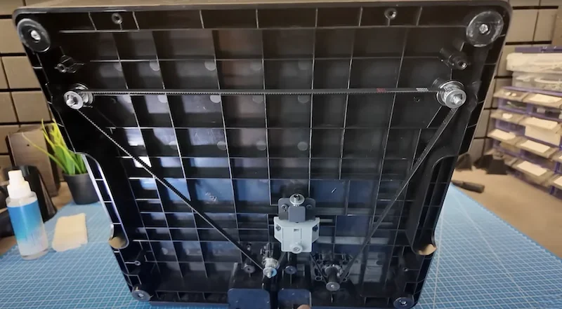 Bottom view of Flashforge Adventurer 5M showing the grid-like structure with belts and pulleys, set on a blue grid mat in a workshop environment.