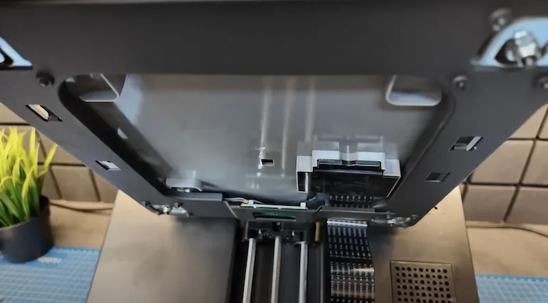 Flashforge Adventurer 5M 3D printer's internal view with visible components, set on a blue mat, plants and tiles in the background.