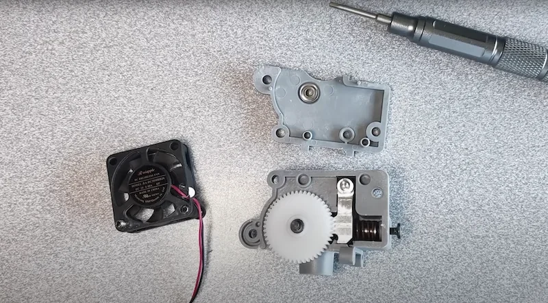 Disassembled parts from Flashforge Adventurer 5M, including a fan, gear mechanism, and screwdriver on a gray surface.