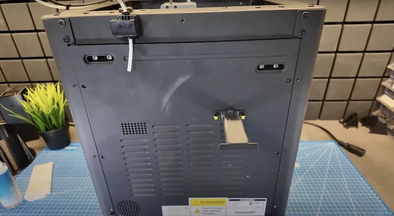 Flashforge Adventurer 5M 3D printer rear view showing ventilation slots, control buttons, and workspace with grid mat and plant.