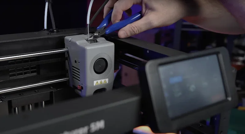 Flashforge Adventurer 5M 3D printer with pliers adjusting filament, touchscreen visible, creating precise 3D parts in a tech workshop setting.