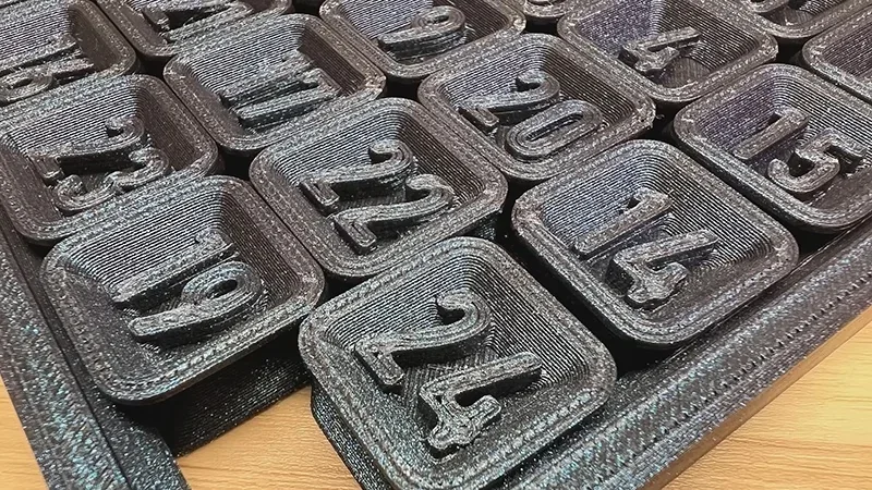 3D-printed numbered black squares on a wooden surface, created by Flashforge Adventurer 5M Pro printer, arranged in rows showing detail.