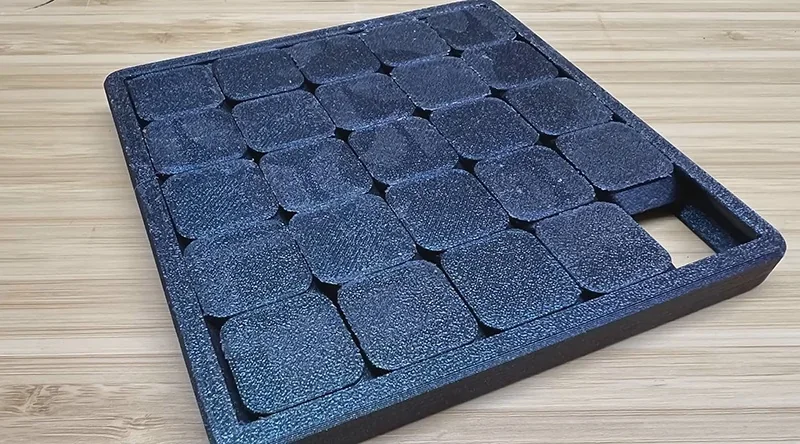 Black grid-style item created by Flashforge Adventurer 5M Pro on a wooden surface; textured squares visible in a rectangular layout.