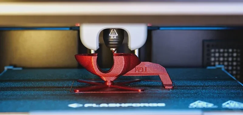 Flashforge Adventurer 5M Pro 3D printer creating a red model on a dark platform with visible branding elements and nozzle head details.