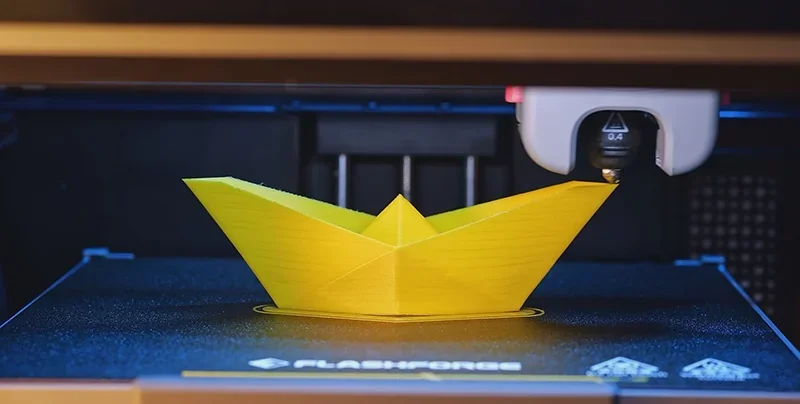 Flashforge Adventurer 5M Pro 3D printer creating a yellow origami-style boat, showcasing precise details on a branded build platform.