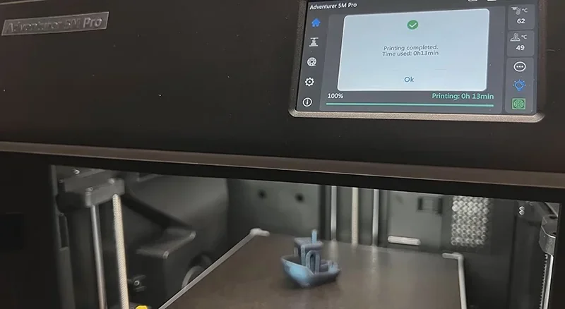 Flashforge Adventurer 5M Pro 3D printer with a completed small boat model on the print bed, screen showing printing status and time, inside a workshop.