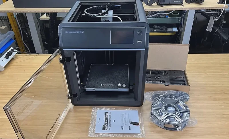 Flashforge Adventurer 5M Pro 3D printer on a table with open door, spool of filament, instruction manual, and accessory kit.