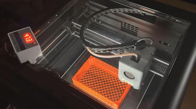 Flashforge Adventurer 5M Pro 3D printer creating an orange lattice container, visible control box showing temperature, sleek black design.