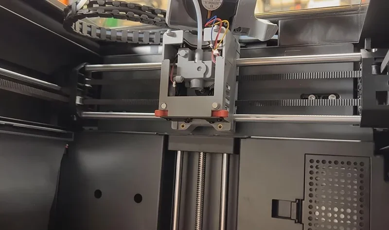 Flashforge Adventurer 5M Pro 3D printer close-up showing extruder assembly, metal rails, and wiring inside a sleek black enclosure.