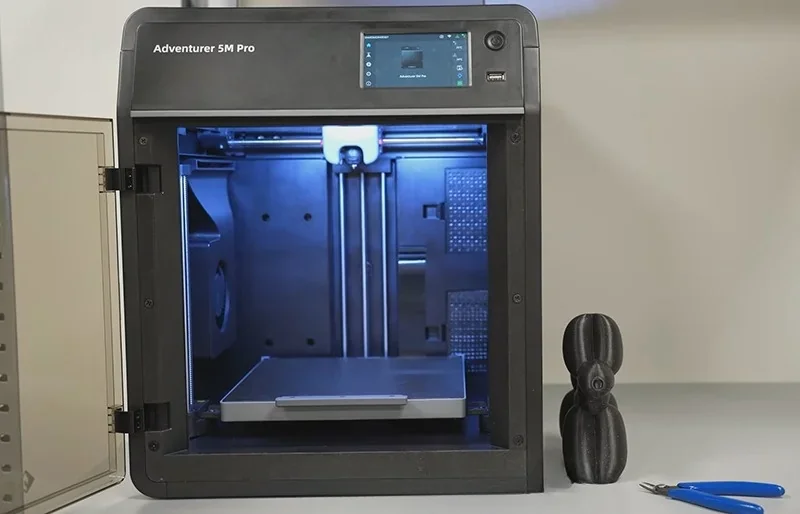 Flashforge Adventurer 5M Pro 3D printer with open build chamber, blue lighting, finished black print model beside unit, tools on a nearby surface.