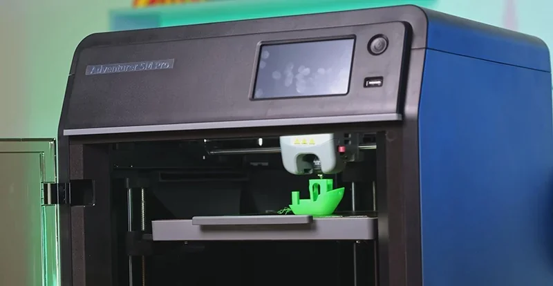 Flashforge Adventurer 5M Pro 3D printer with touchscreen, printing a green model boat inside its black and blue enclosure.