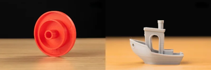 Flashforge Creator 4 output: a red circular component and a gray 3D-printed boat on a wooden surface, showcasing precise detailing.