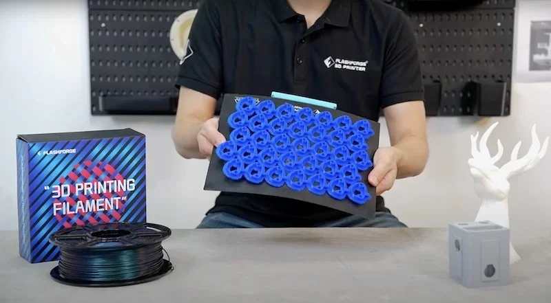 FlashForge Guider 3 Ultra showing blue 3D printed parts on a tray, filament spool, and white figurine, with branded packaging in a workshop setting.