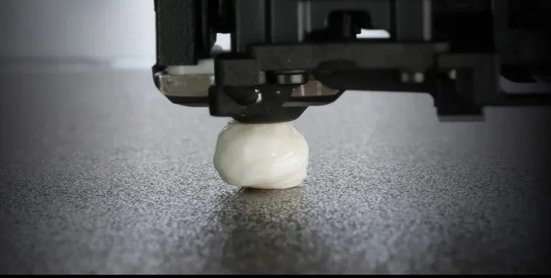 H2c Bambu 3D printer nozzle printing a small white object on a textured surface, highlighting precision and technology.