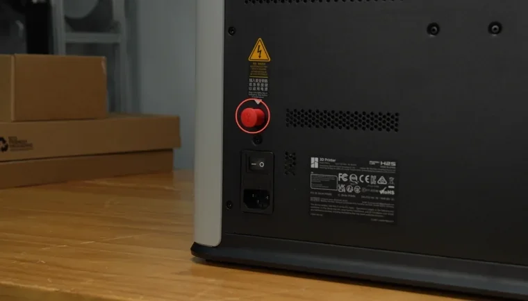 Bambu 3D printer rear view with warning label, red knob, power switch; cardboard boxes in background.