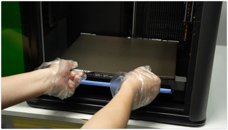 Hands in gloves handling h2c Bambu 3D printer bed, focusing on precise removal, showcasing advanced technology and craftsmanship.