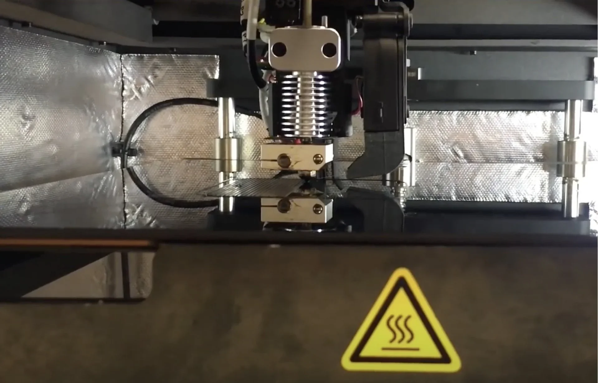 Intamsys Funmat HT 3D printer in action, extruder with a reflective enclosure and caution symbol, creating detailed parts.
