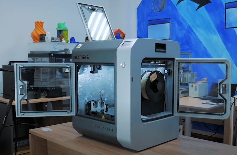 Intamsys Funmat HT 3D printer with open doors on a desk; colorful 3D-printed samples in background; modern studio setting.