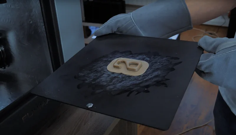 Gloved hands hold a 3D printed part on a black plate from the Intamsys Funmat HT printer, with branding visible, in a workshop setting.