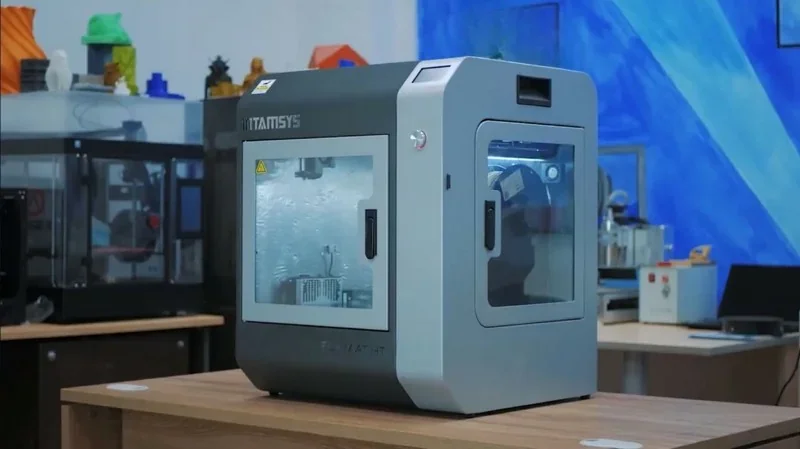 Intamsys Funmat HT 3D printer in a workshop setting, showcasing its sleek design and transparent door with 3D printed samples visible in the background.