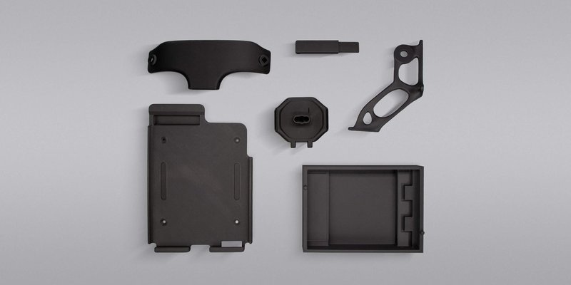 Black 3D-printed parts created by the Intamsys Funmat Pro 310 are arranged against a gray background. The parts include a rectangular housing with screw holes, an L-shaped component with geometric cutouts, and other precision-engineered pieces. Each item showcases the printer's capability for producing detailed and varied shapes in a sleek, matte finish.