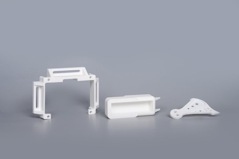 A set of three white 3D printed components created by the Intamsys Funmat Pro 310. The parts include structural pieces and an intricate bracket design, exemplifying the printer's capability to produce complex and precise geometries. The objects are displayed against a smooth, neutral backdrop, focusing attention on their detailed craftsmanship.
